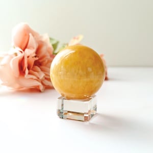May include: A polished, golden-yellow sphere, possibly a gemstone or decorative object, sits atop a clear, square crystal base. Soft pink flowers are blurred in the background, creating a delicate contrast. The sphere's smooth surface reflects light.