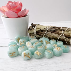May include: A set of 20 teal green tumbled stones with gold runes inscribed on each. The stones are arranged in a pile on a white surface. A bundle of dried sage is in the background.