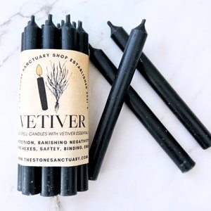 May include: A bundle of black Vetiver spell candles with a brown label. The label features the text "VETIVER" and "THE STONE SANCTUARY". The candles are for protection, banishing negativity, and binding. The candles are approximately 5 inches long.