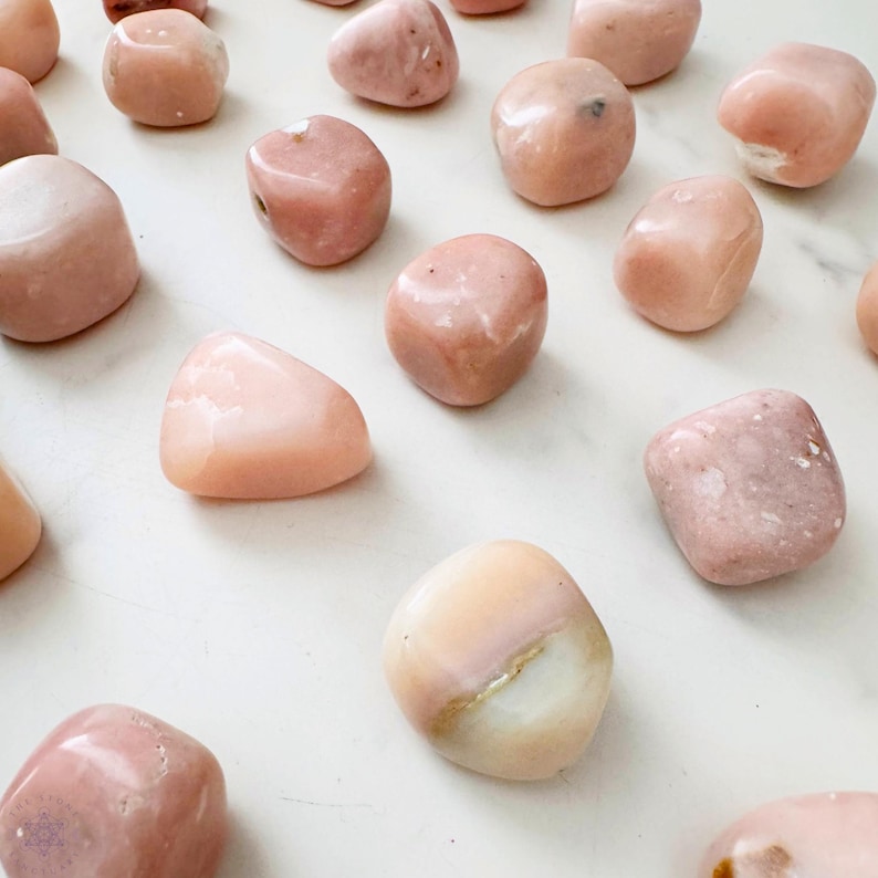 May include: A collection of polished pink opal stones, each with a smooth, rounded shape. The stones vary in shade from pale to deeper pink, with some showing a mix of colors. The arrangement is scattered across a white surface.