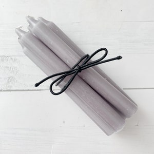 May include: A bundle of six lavender-colored taper candles tied together with a black elastic cord. The candles are arranged diagonally, with their wicks visible. The background is a white wooden surface.