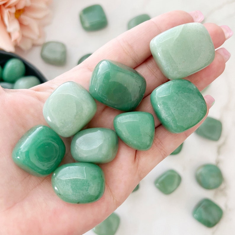 May include: A collection of smooth, green tumbled stones. The stones are various shades of green, from light to dark. They are arranged in a hand, with some stones spilling out onto a white surface.