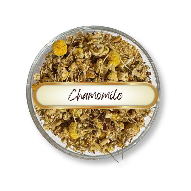 May include: A clear glass bowl filled with dried chamomile flowers and buds. The bowl has a gold-edged label with the word "Chamomile" in a script font. The chamomile is a mix of yellow and brown hues.