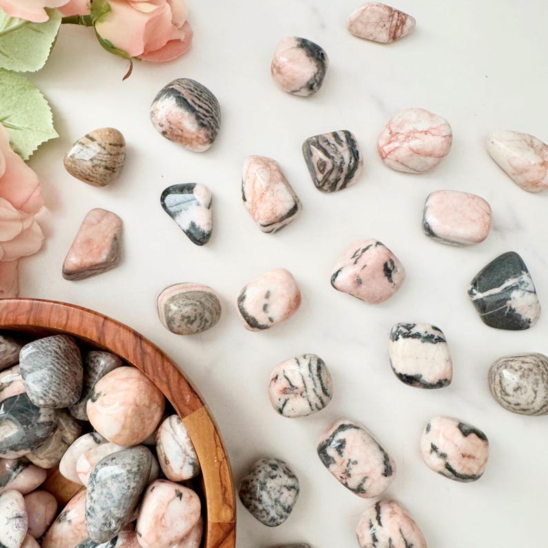 May include: A collection of polished stones in shades of pink, gray, and black, scattered on a white surface. Some stones have striped patterns, while others are solid. A wooden bowl filled with similar stones is visible.