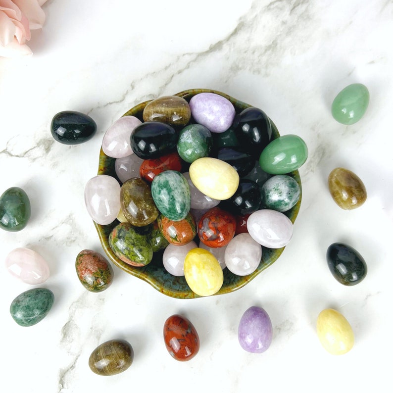 Puede incluir: Un bol lleno de huevos de piedras preciosas pulidas de varios colores. Los huevos son en tonos de verde, rosa, rojo, amarillo, morado y negro. El bol es verde y marr&oacute;n con una superficie texturizada.