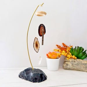 May include: A gold metal stand with a black agate base holds a crystal wind chime with three agate slices and a clear quartz crystal. The wind chime hangs from a wooden disc.