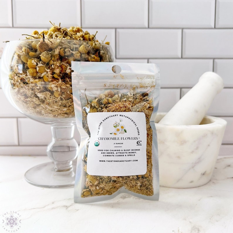 May include: A clear glass bowl and a clear bag filled with dried chamomile flowers. The bag has a label that reads "Chamomile Flowers" and lists uses. A white mortar and pestle are on the right.