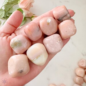 May include: A handful of polished, tumbled pink stones. The stones vary in shade from light to medium pink, with some showing white and gray inclusions. The stones are smooth and rounded, and the image is taken in natural light.
