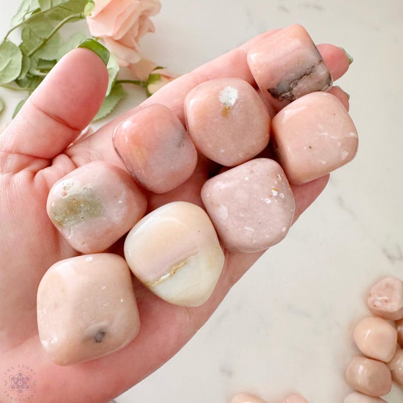 May include: A handful of polished, tumbled pink stones. The stones vary in shade from light to medium pink, with some showing white and gray inclusions. The stones are smooth and rounded, and the image is taken in natural light.
