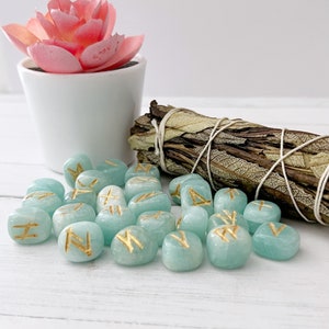 May include: A set of 24 teal green gemstone runes with gold lettering. The runes are scattered on a white surface next to a bundle of dried sage.