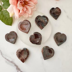 May include: A collection of heart-shaped stone dishes in varying shades of brown and red, with subtle green and blue veining. The dishes are arranged on a white surface, with a pink flower and green leaves in the background.