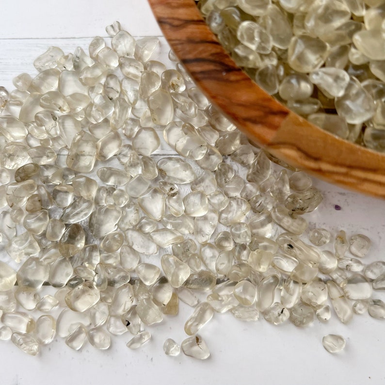 Puede incluir: Primer plano de un mont&oacute;n de piedras de cuarzo transparente pulidas. Las piedras son lisas y tienen una apariencia natural y transl&uacute;cida. Est&aacute;n esparcidas sobre una superficie blanca.
