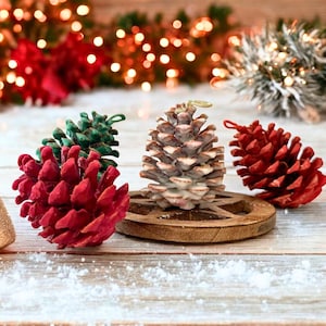 May include: Four pine cones, one green, one red, one white, and one red, on a wooden surface. The pine cones are decorated for the holidays.