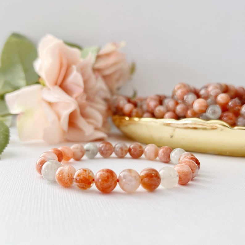 May include: A bracelet made of orange, white, and red sunstone beads. The bracelet is on a white surface with a pink rose in the background.