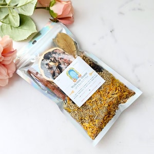 May include: A clear, iridescent pouch containing a simmer pot mix with dried orange slices, cinnamon sticks, bay leaves, and yellow flower petals. A white label reads Welcome Home SIMMER POT MIX, A WELCOMING SIMMER POT BLEND TO BLESS, PROTECT & SETTLE A NEW HOME.