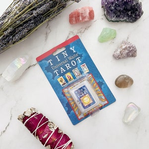 May include: A packaged "TINY Universal Waite Tarot" card deck with a blue and red card. The image also features various crystals, a bundle of dried lavender, and a sage bundle wrapped with rose petals.