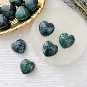 May include: A collection of polished heart-shaped stones in shades of green and dark teal. Some hearts are on a gold-colored tray, others on a white circular surface. A sprig of lavender is visible in the background.