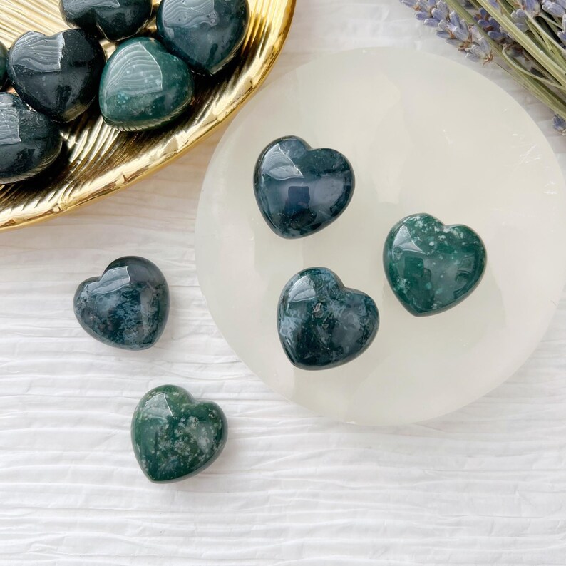 May include: A collection of polished heart-shaped stones in shades of green and dark teal. Some hearts are on a gold-colored tray, others on a white circular surface. A sprig of lavender is visible in the background.