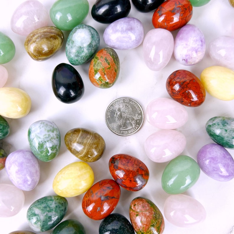Puede incluir: Una colecci&oacute;n de huevos de piedras preciosas pulidas en varios colores, incluyendo verde, morado, rosa, rojo, marr&oacute;n y amarillo. Los huevos est&aacute;n dispuestos en un c&iacute;rculo alrededor de una moneda de veinticinco centavos de d&oacute;lar estadounidense.