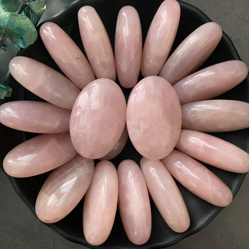 Pu&ograve; includere: Una collezione di 15 pietre di quarzo rosa lisce e levigate di diverse dimensioni e forme. Le pietre sono disposte in un cerchio su una superficie nera.