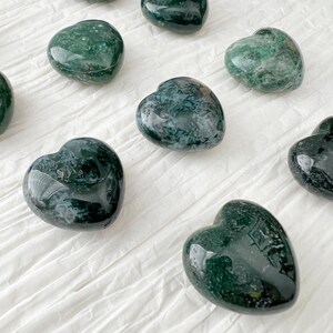 May include: A collection of polished heart-shaped stones. The stones are a deep green color with lighter green and white marbling. They are arranged on a white, textured surface.