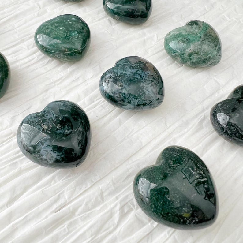 May include: A collection of polished heart-shaped stones. The stones are a deep green color with lighter green and white marbling. They are arranged on a white, textured surface.