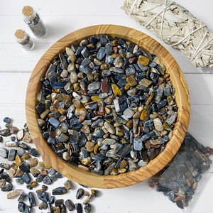May include: A wooden bowl filled with a variety of blue and gold tiger's eye stones. The stones are small and irregular in shape, and they are arranged in a random pattern. The bowl is sitting on a white surface.