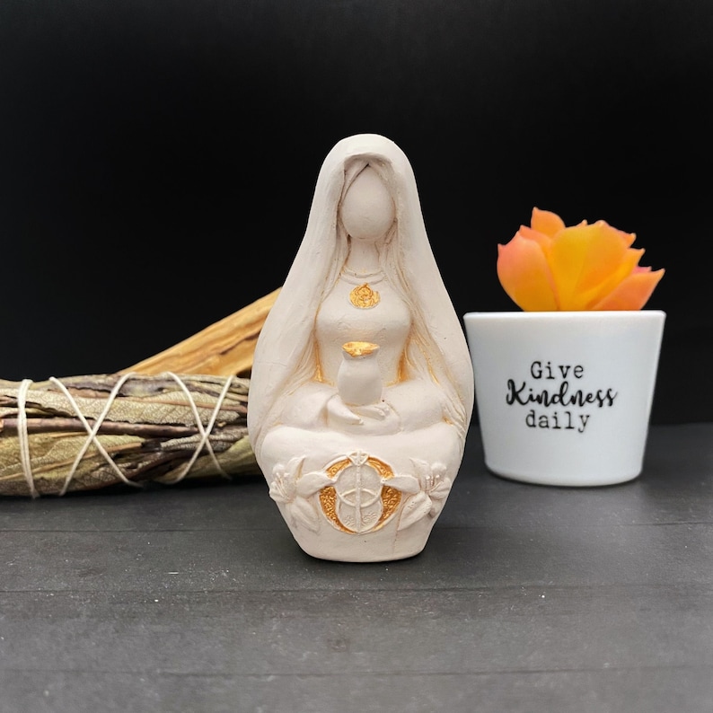 Puede incluir: Una estatua de color crema de una figura sentada con la cabeza cubierta con una tela. La figura sostiene un peque&ntilde;o recipiente y est&aacute; adornada con detalles dorados. A la derecha hay una peque&ntilde;a maceta blanca con el texto "Give Kindness daily" y una flor naranja artificial.