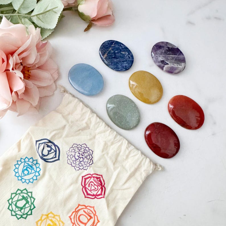 May include: A collection of seven polished chakra stones in various colors, including blue, purple, yellow, and red, arranged on a white surface. A beige cloth bag with chakra symbols is also present. Pink flowers are in the background.