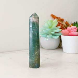 May include: A tall, green and white crystal obelisk with a pointed top. The polished stone has a natural, marbled appearance. The obelisk is standing upright. The background includes a white surface and decorative plants in white pots.