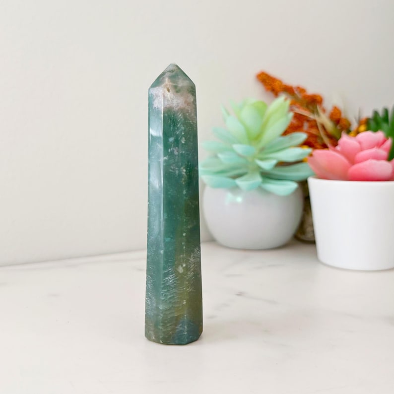 May include: A tall, green and white crystal obelisk with a pointed top. The polished stone has a natural, marbled appearance. The obelisk is standing upright. The background includes a white surface and decorative plants in white pots.