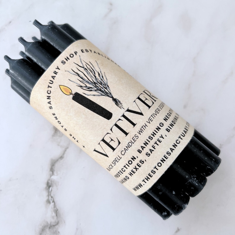 May include: A bundle of black spell candles wrapped in a tan paper label. The label features the word "VETIVER" in large letters, along with text about protection and banishing negativity. The Stone Sanctuary Shop is also printed on the label.