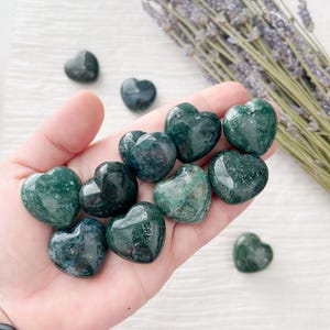 May include: A collection of polished green moss agate heart-shaped stones. The stones vary in shade from deep forest green to lighter hues, with visible patterns. A sprig of dried lavender is in the background.