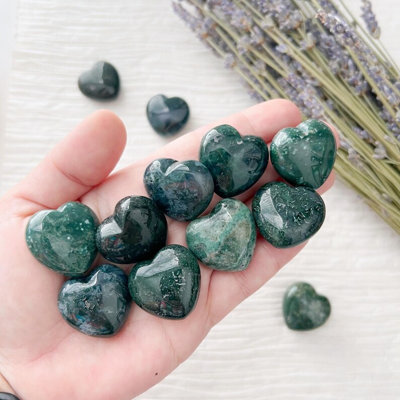 May include: A collection of polished green moss agate heart-shaped stones. The stones vary in shade from deep forest green to lighter hues, with visible patterns. A sprig of dried lavender is in the background.