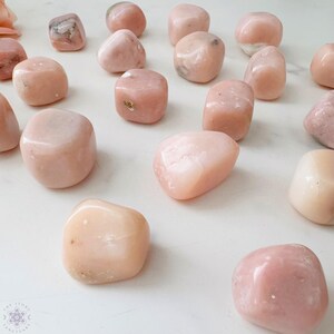 May include: A collection of smooth, polished pink opal stones. The stones vary in shape and size, with a soft, translucent appearance. The background is a clean, white surface, highlighting the natural beauty of the gemstones. The stones are scattered across the surface.