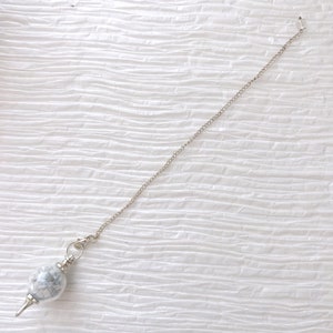 May include: A silver chain with a white and gray stone pendulum. The pendulum is round and has a pointed tip.