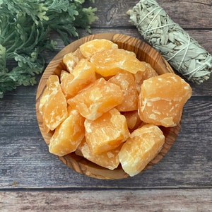 May include: A wooden bowl filled with raw, orange calcite crystals. The crystals have a rough, natural texture and vary in size. A bundle of white sage is visible in the background, along with green foliage. The bowl sits on a wooden surface.