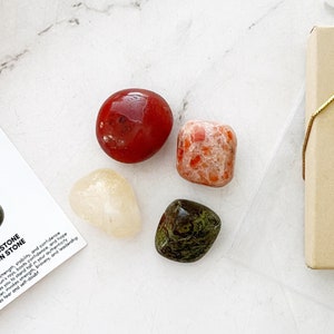 May include: Set of four tumbled stones: red carnelian, orange sunstone, clear citrine, and green aventurine. The stones are arranged on a white surface with a small brown box in the background.