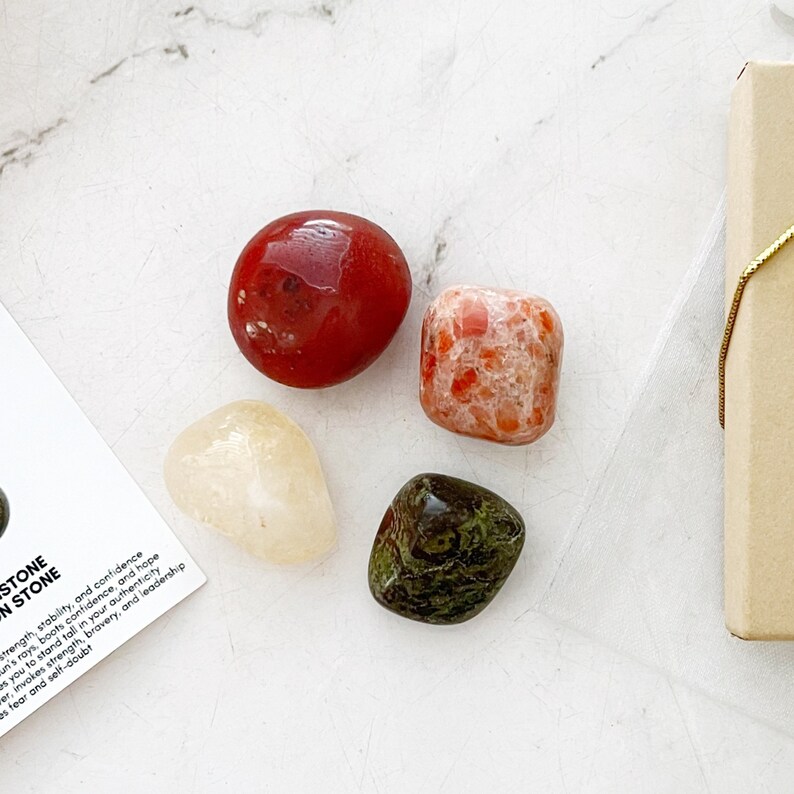 May include: Set of four tumbled stones: red carnelian, orange sunstone, clear citrine, and green aventurine. The stones are arranged on a white surface with a small brown box in the background.