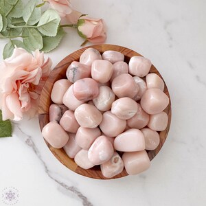 May include: A wooden bowl filled with smooth, polished pink stones. The stones vary in shade from pale to deeper pink, with some showing subtle veining. Soft pink flowers and green leaves are arranged near the bowl on a white marble surface.