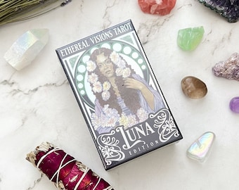 May include: A tarot card deck titled "Ethereal Visions Tarot Luna Edition" featuring a woman with floral accents. The image includes crystals, sage, and lavender on a marble surface.
