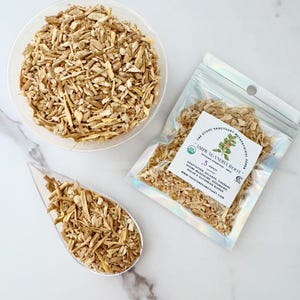 May include: An overhead shot displays ashwagandha root in a clear bowl, a clear scoop, and a sealed bag. The bag has a label that reads "ASHWAGANDHA ROOT" and "5 ounces". The root pieces are light brown and appear to be dried.
