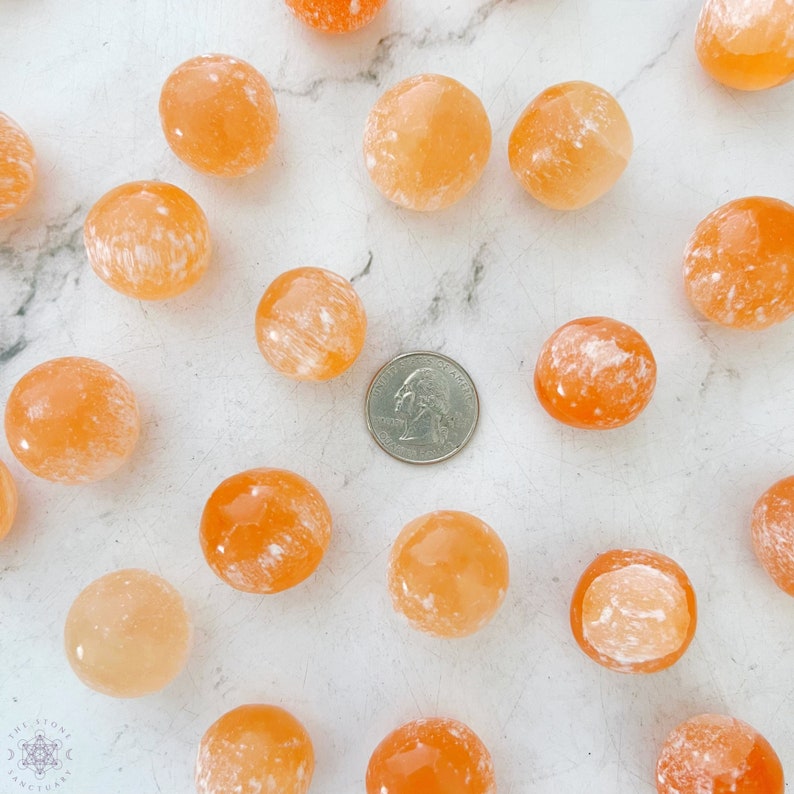 May include: A collection of 18 orange and white tumbled carnelian stones. The stones are various sizes and shapes, and some have a more pronounced white pattern than others. A US quarter is included for size reference.