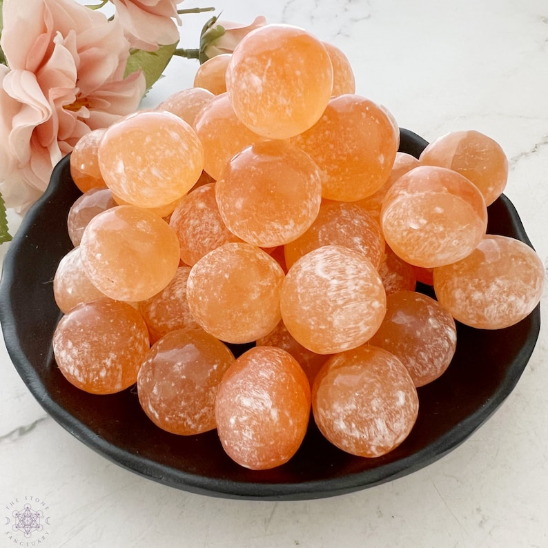 May include: A collection of orange carnelian tumbled stones in a black bowl. The stones are smooth and have a natural, polished finish.
