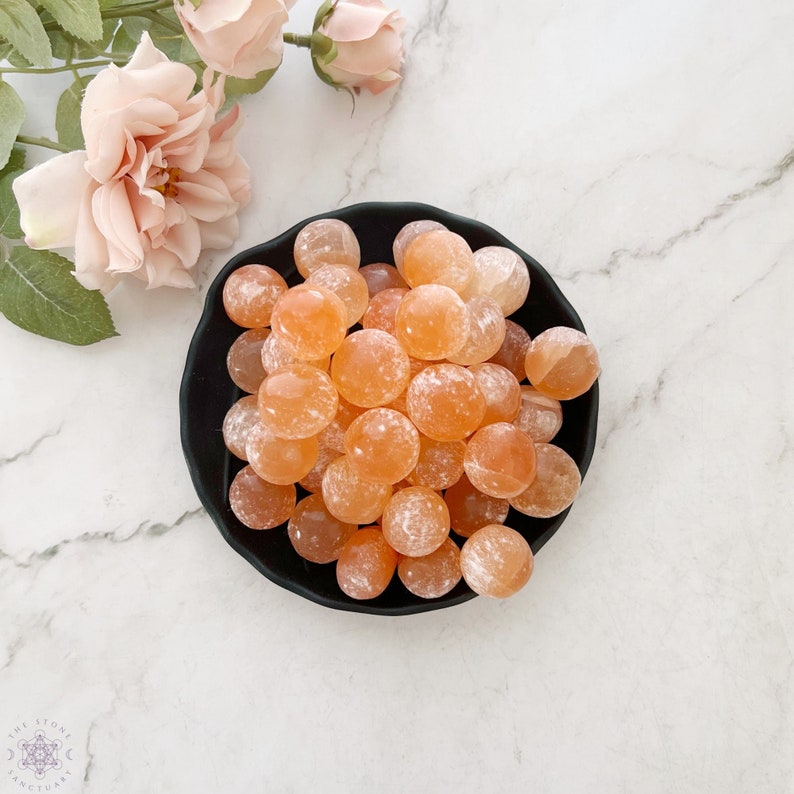 May include: A black bowl filled with approximately 25 orange and white carnelian tumbled stones. The stones are smooth and polished.