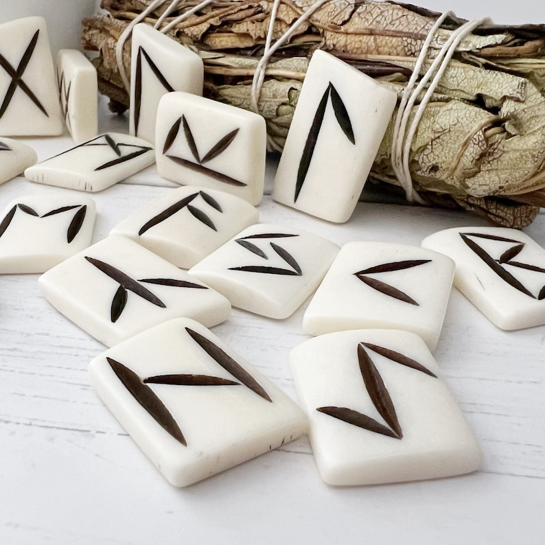 May include: Ivory-colored rune stones with dark brown markings, arranged on a white surface. A bundle of dried sage is in the background. The runes are rectangular and feature various symbols.