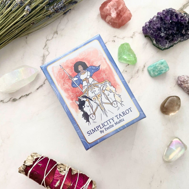May include: A tarot card deck titled "Simplicity Tarot" by Emilie Mu&ntilde;iz, featuring an illustration of a woman riding two horses. The deck is surrounded by crystals, dried lavender, and a sage bundle, all arranged on a white marble surface.