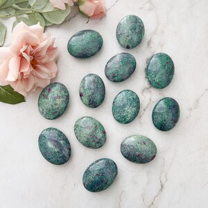 May include: A collection of green and red oval-shaped tumbled stones. The stones have a speckled appearance and are arranged on a white marble surface.