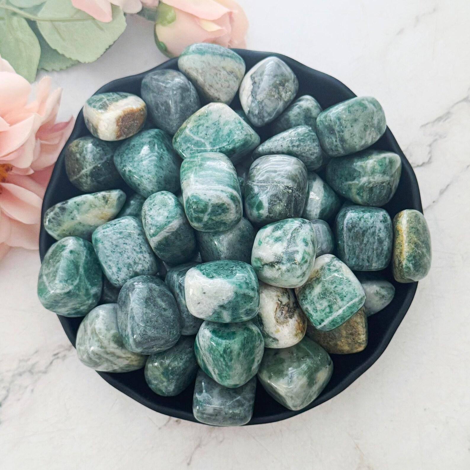 Tumbled green jade stones in a black bowl on marble, surrounded by soft pink flowers.