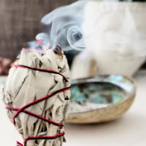 May include: White sage smudge stick tied with red string, burning and releasing smoke. The sage is in a white abalone shell.